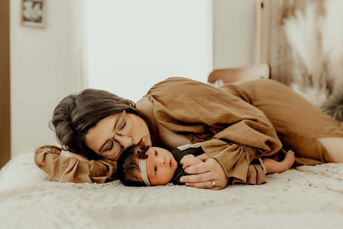 A woman with glasses lies on a bed, gently embracing a baby wearing a headband. The room is softly lit.