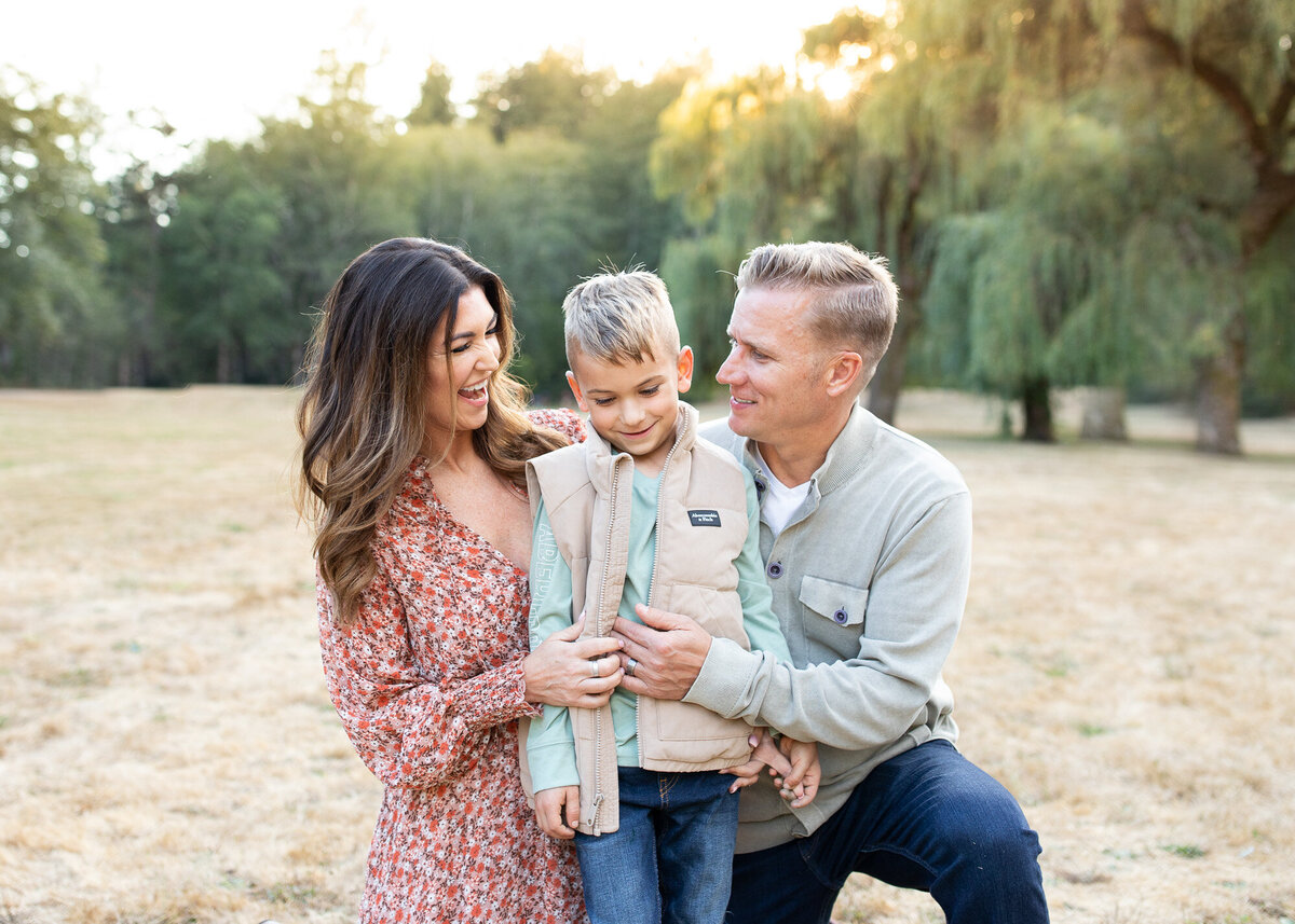 Family of three kneeling in grass at sunset with large trees behind them. Photo by Portland Or family portrait photography.