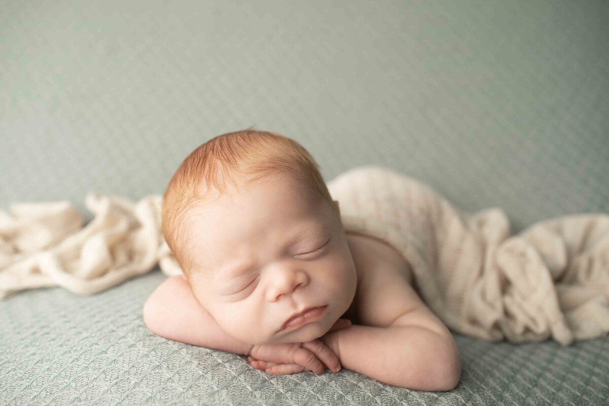 Newborn baby boy with vibrant red hair posed on a soft backdrop, his tiny feet tucked close in a cozy position
