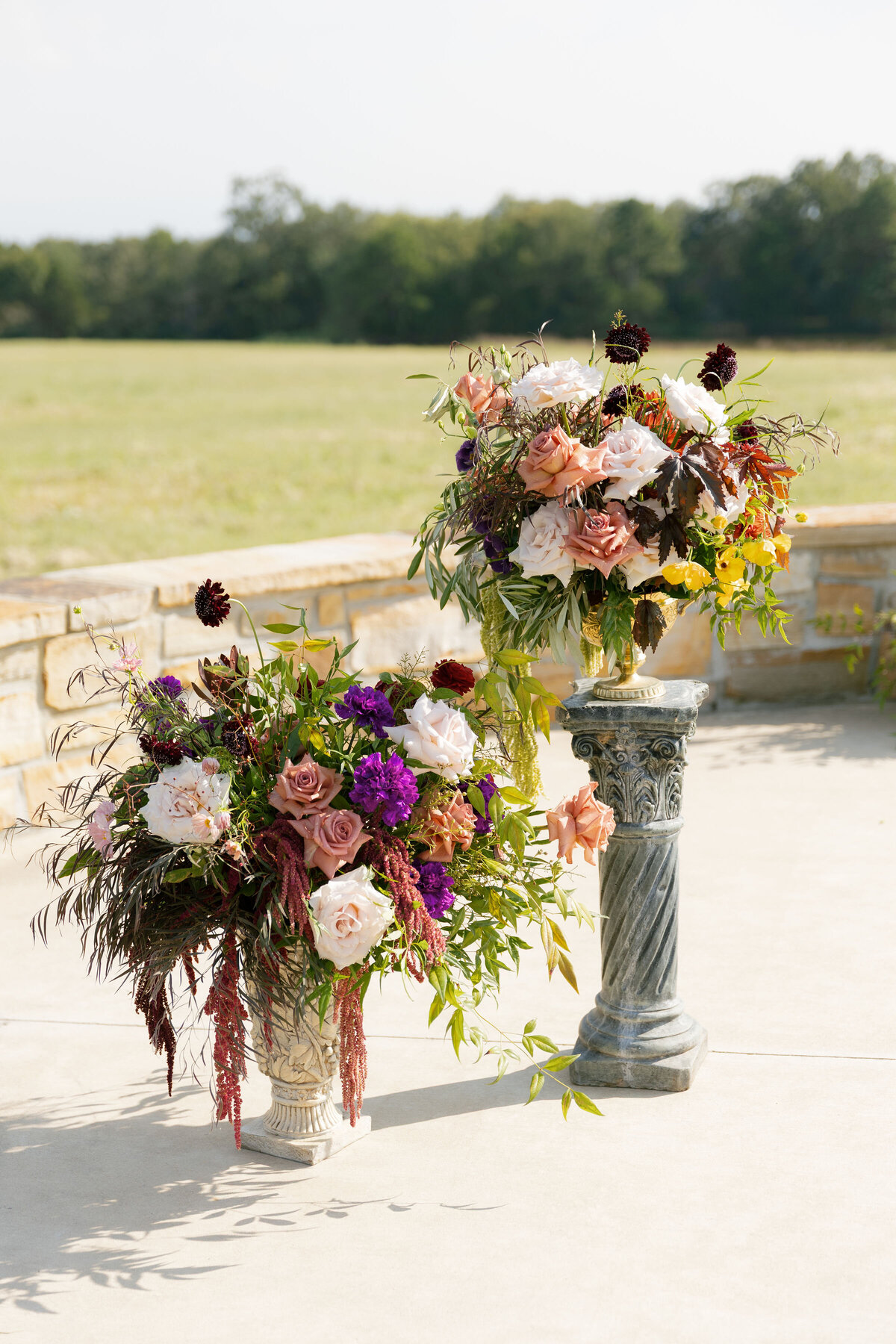 Two elaborate floral arrangements sit atop ornate pedestal stands for an outdoor wedding ceremony setup, overflowing with roses in soft blush, peach, mauve, deep burgundy blooms, purple accent flowers, and cascading greenery. The arrangements frame the ceremony space with lush, garden-inspired color against an open field backdrop at a modern Arkansas venue.