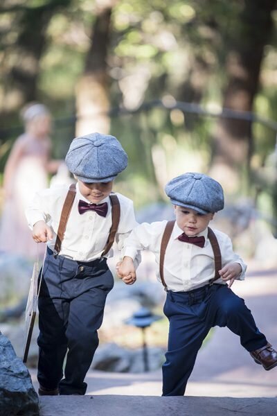 Two small ring bearers walk up the aisle holding hands and wearing hats, suspenders, and bow ties.