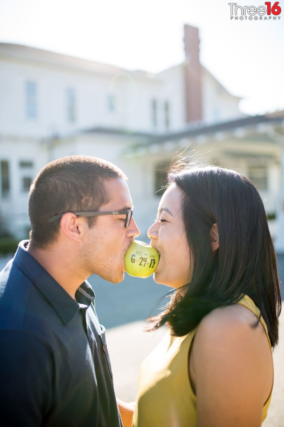 Engaged couple hold an apple together in their teeth's with their wedding date written on it