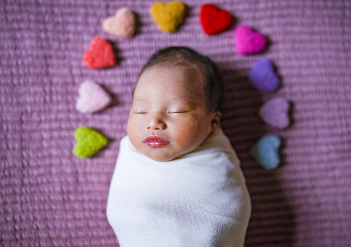 newborn girl laying on purple blanket wrapped up in white blanket with rainbow hearts about her head