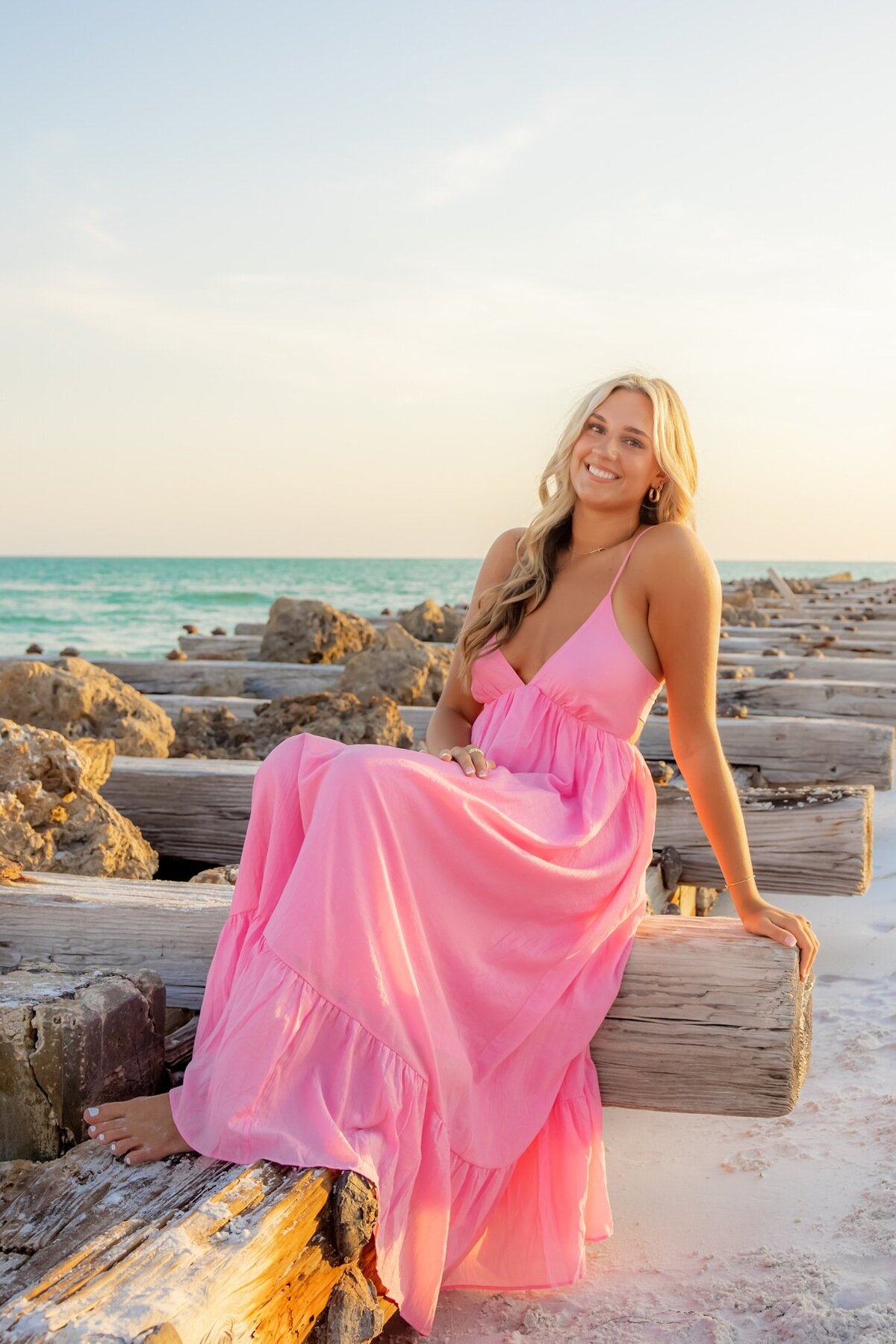 Pretty senior girl in long pink dress poses seated for Love and Style Photography on a jetty in Anna Maria Island.
