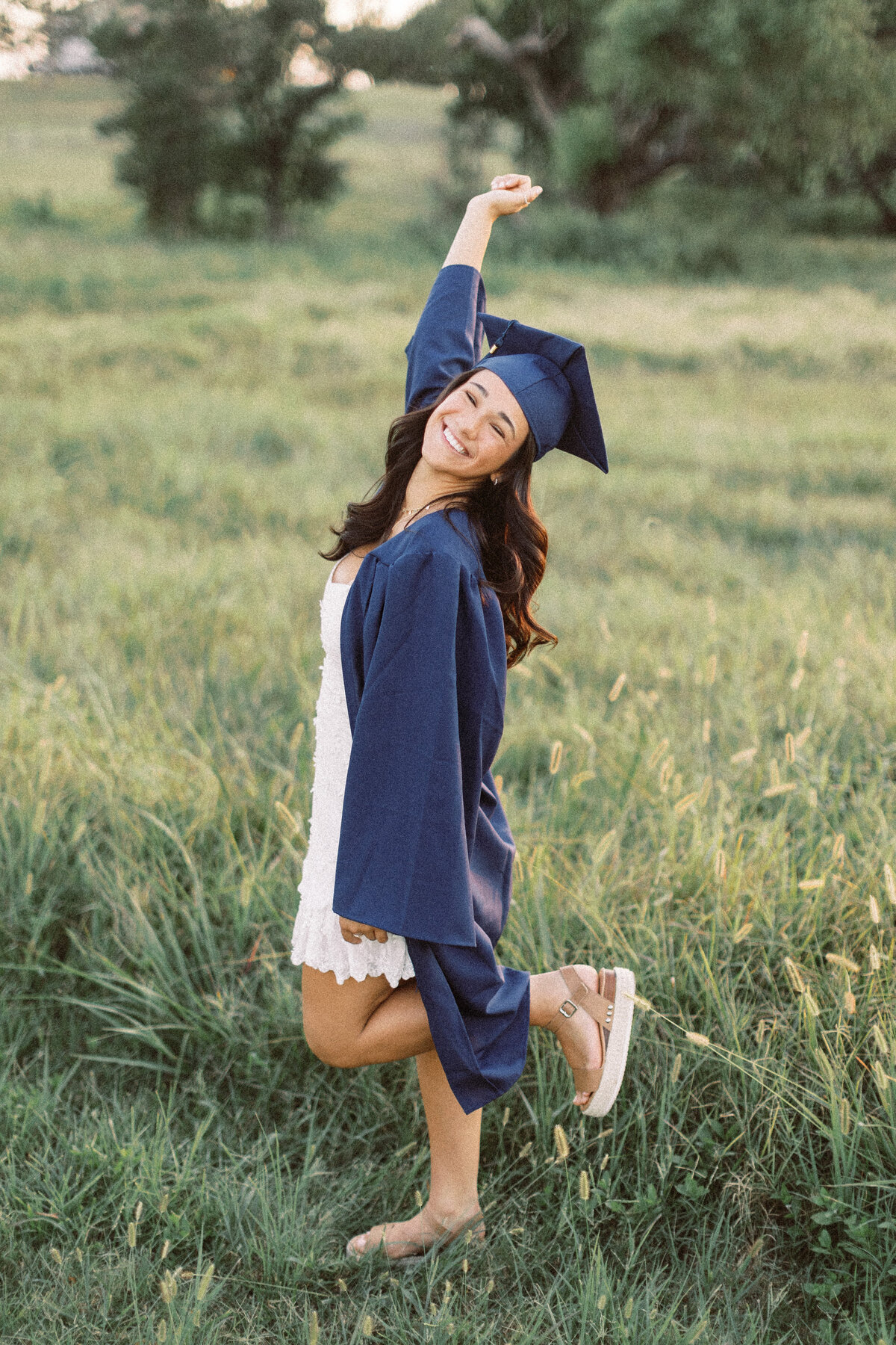 young woman in navy graduation cap and gown celebrates with arm in the air