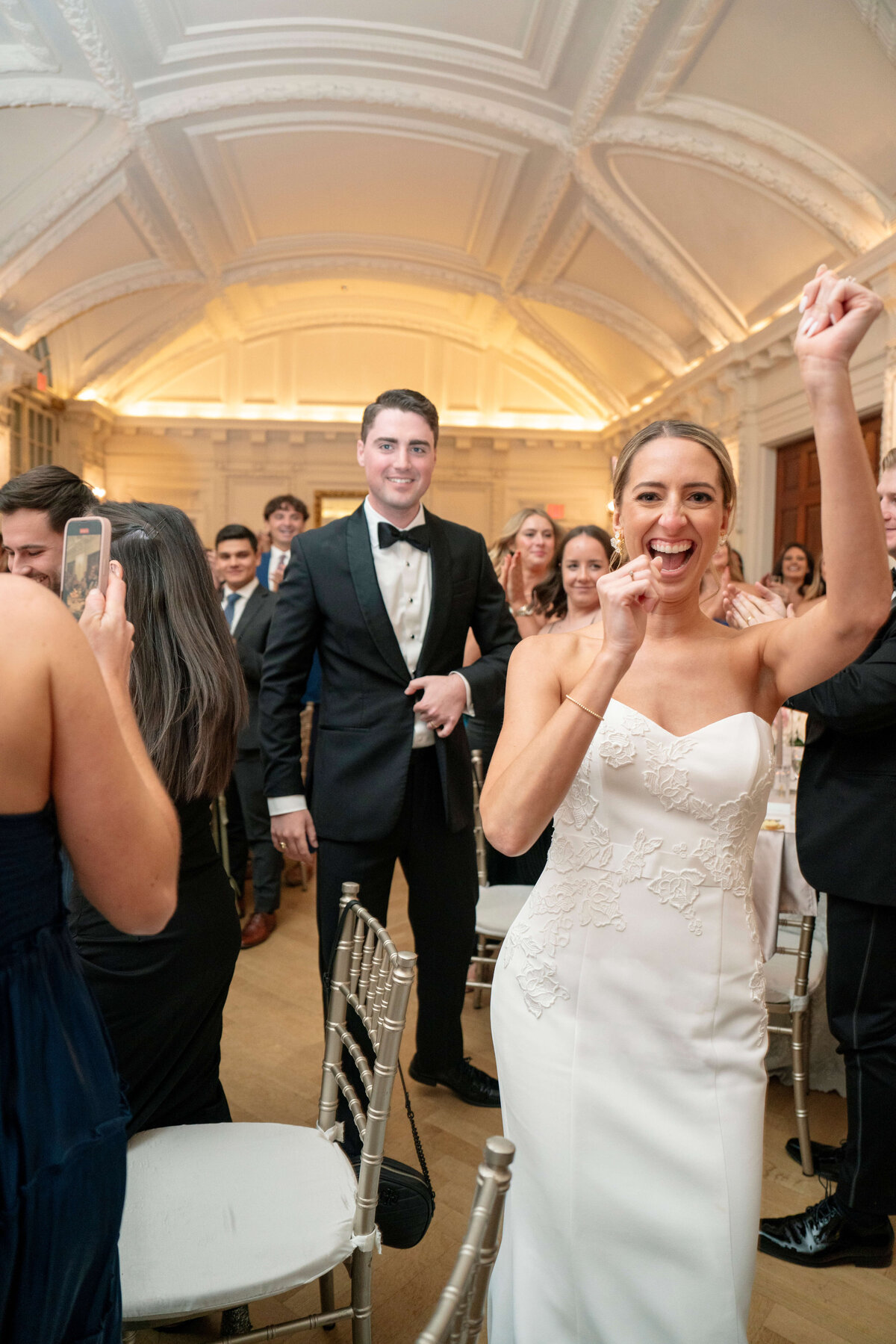 A bride in a white gown joyfully raises her hand while guests celebrate around her. The groom, in a black tuxedo, stands happily in the background. The setting is an elegantly decorated hall with a high, white ceiling.