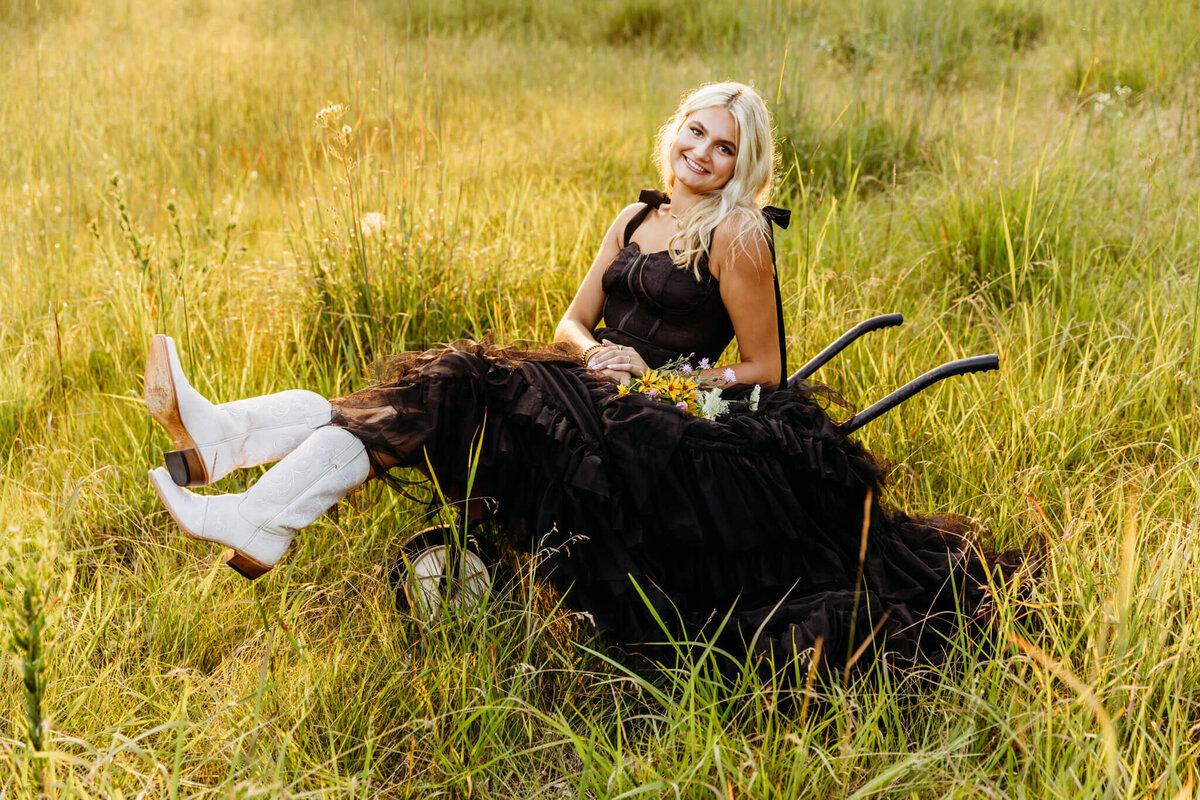 portrait of a teenage girl in a black dress sitting in a wheel barrow with her white cowboy boots handing out the front as she holds a bouquet of flowers captured by Ashley Kalbus Photography