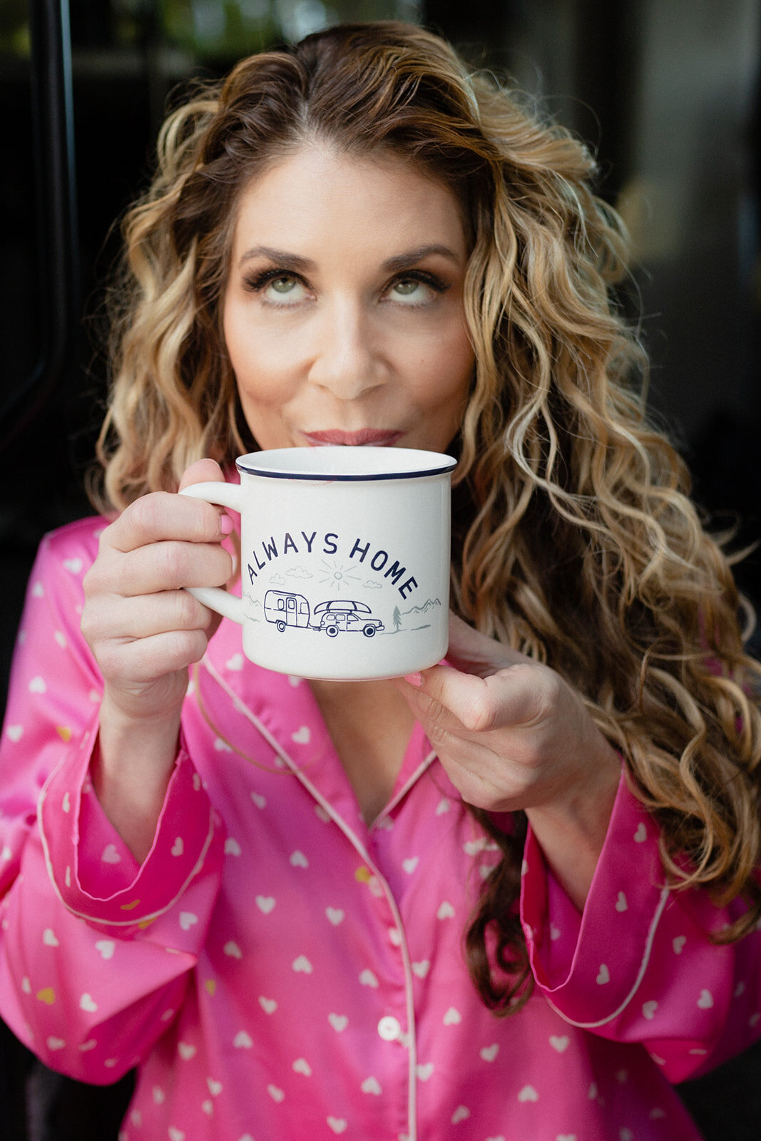 close up shot of a woman sitting in pink pjs with white hearts, drinking from a mug that says Always Home, and looking upward.