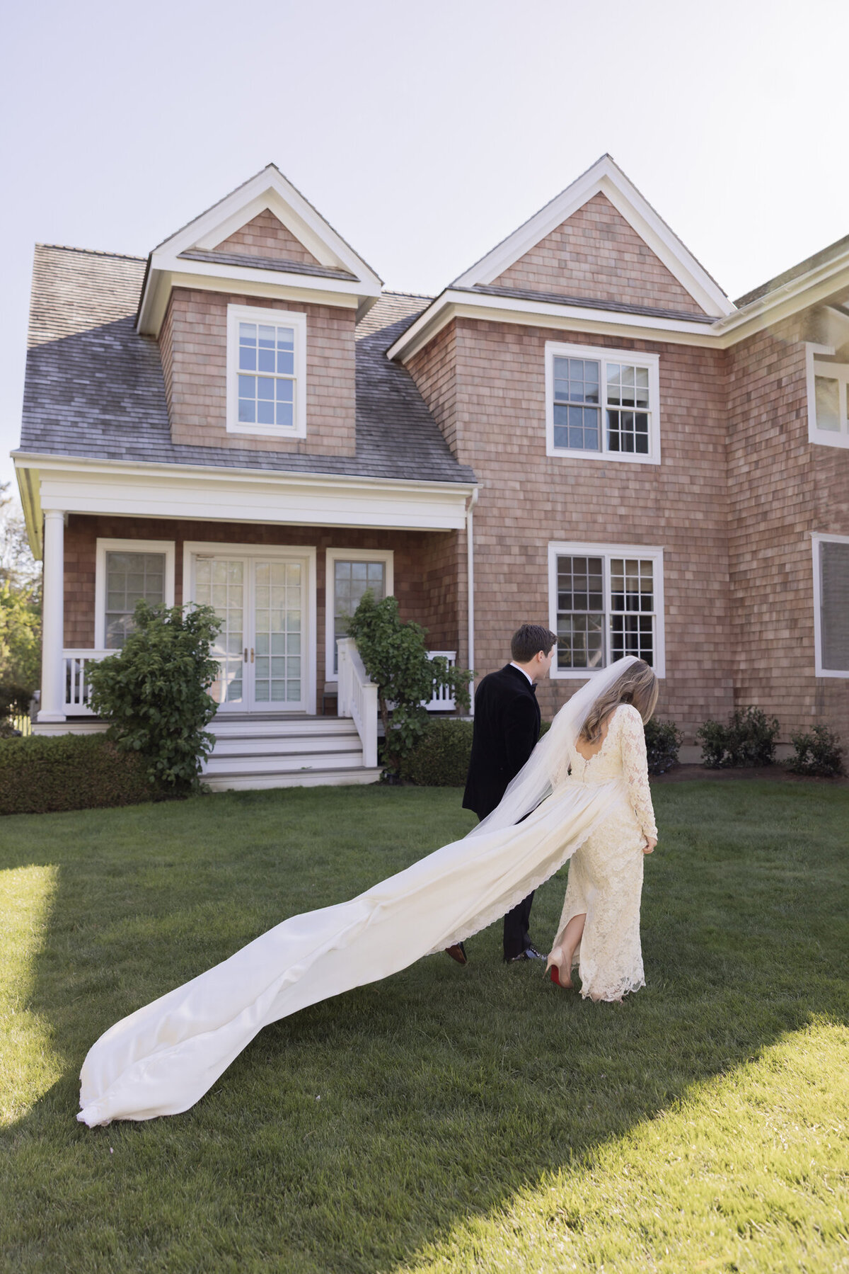 Bride and groom walk hand-in-hand across the lawn of a private Hamptons estate following their outdoor ceremony, with the bride’s long train and veil trailing behind her.