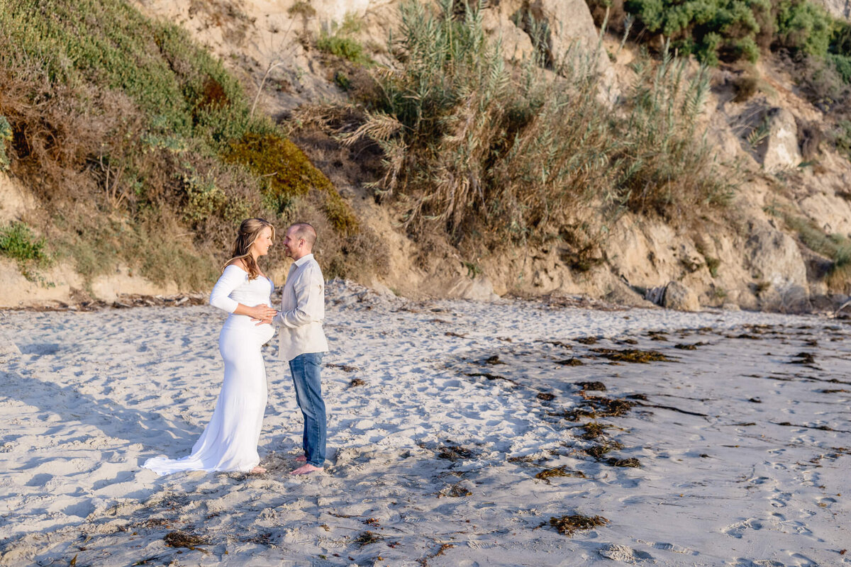 A couple stands on a sandy beach near rocky cliffs, with the woman wearing a long white dress and the man in a light sweater and jeans. They are holding hands and looking at each other, surrounded by natural coastal vegetation.