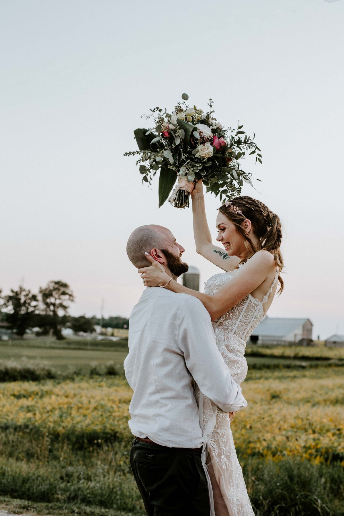 Groom lifting his bride for fun wedding day photo in Exeter, Ontario with barns and pastures behind them. Bride's forearm is holding the groom's neck and her back arm is raising a large bouquet.