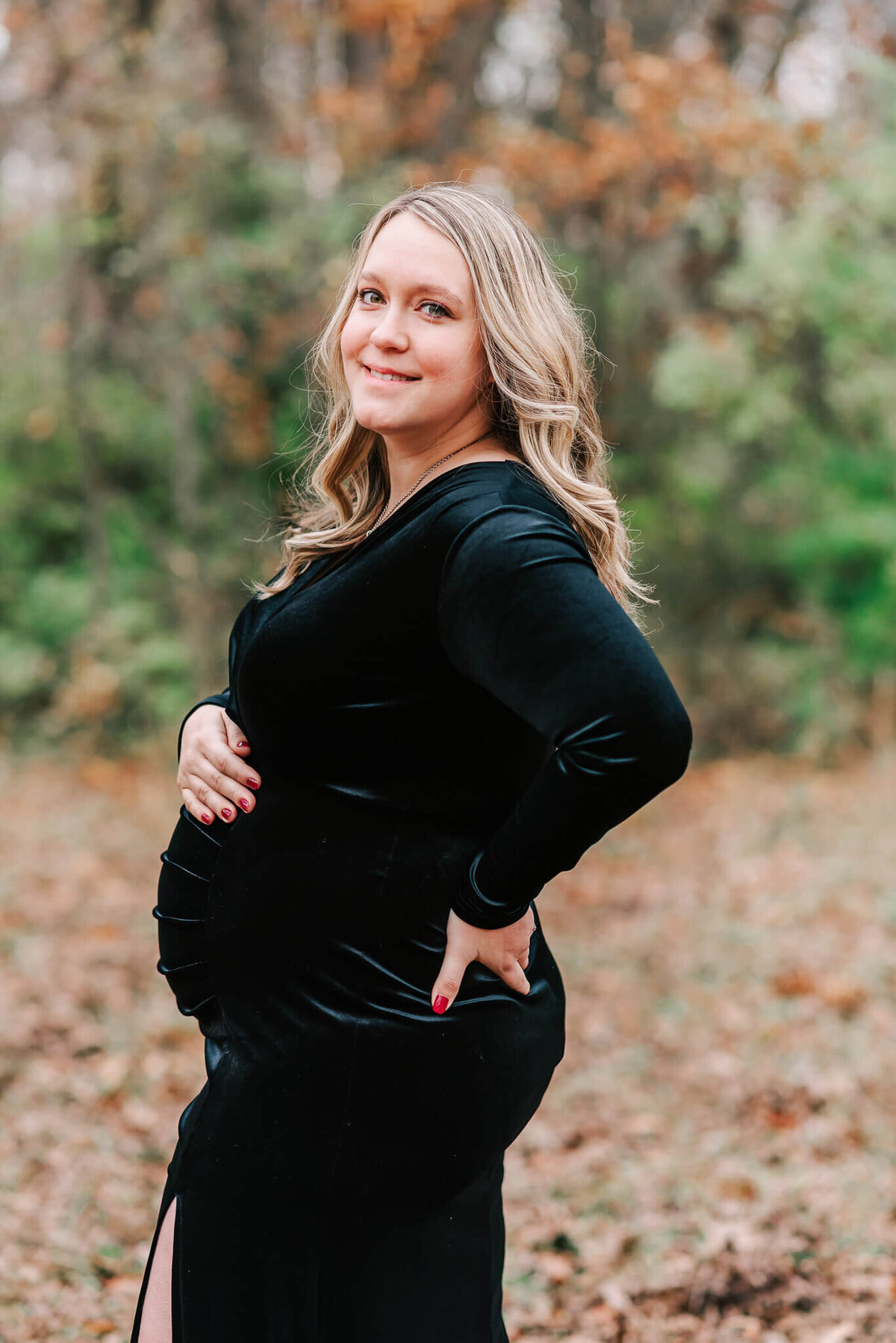 A mama in a floor-length black velvet dress smiling at the camera during her maternity session
