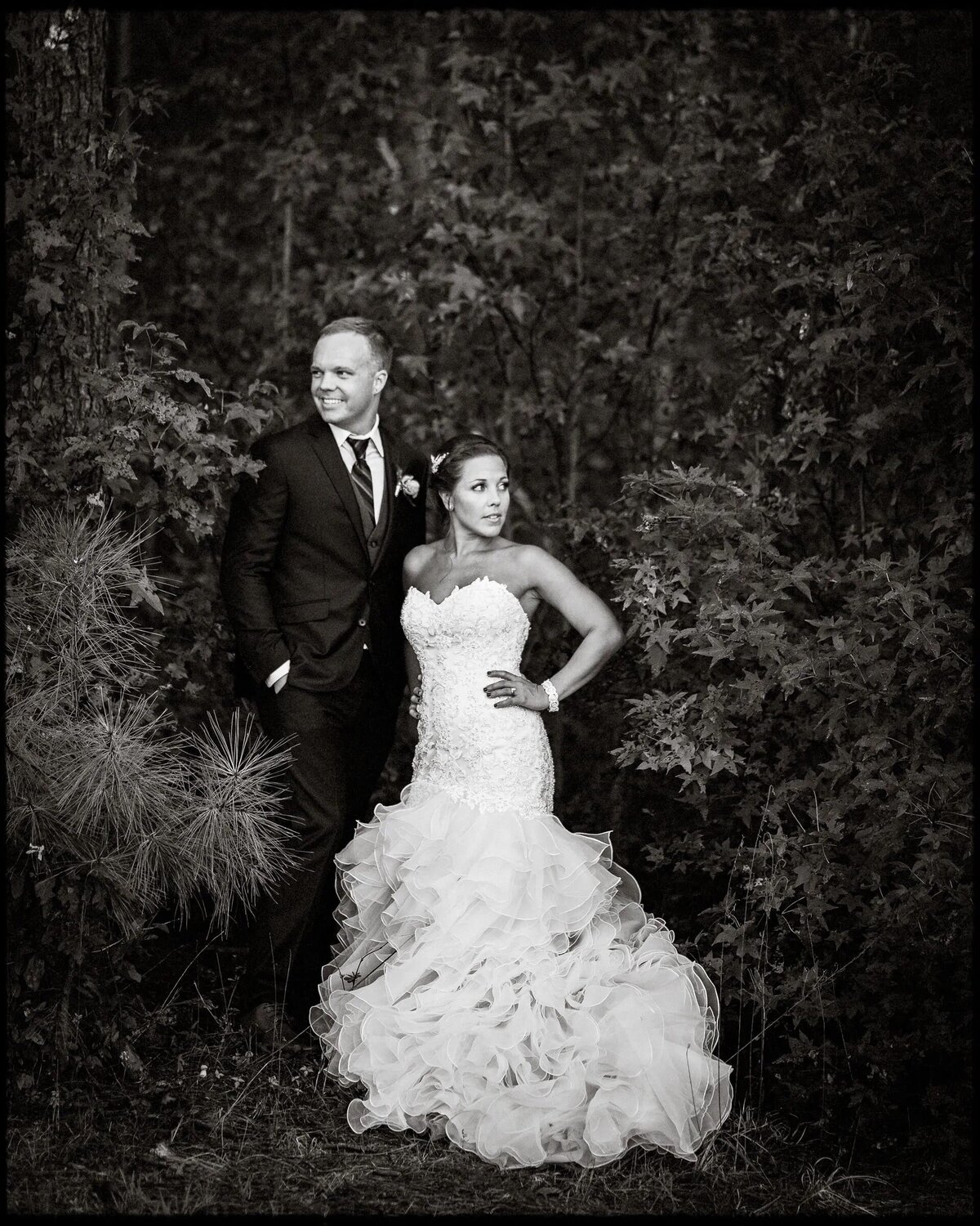 Black and white portrait of a bride and groom in a forest setting, with the groom looking at the camera and the bride looking away.