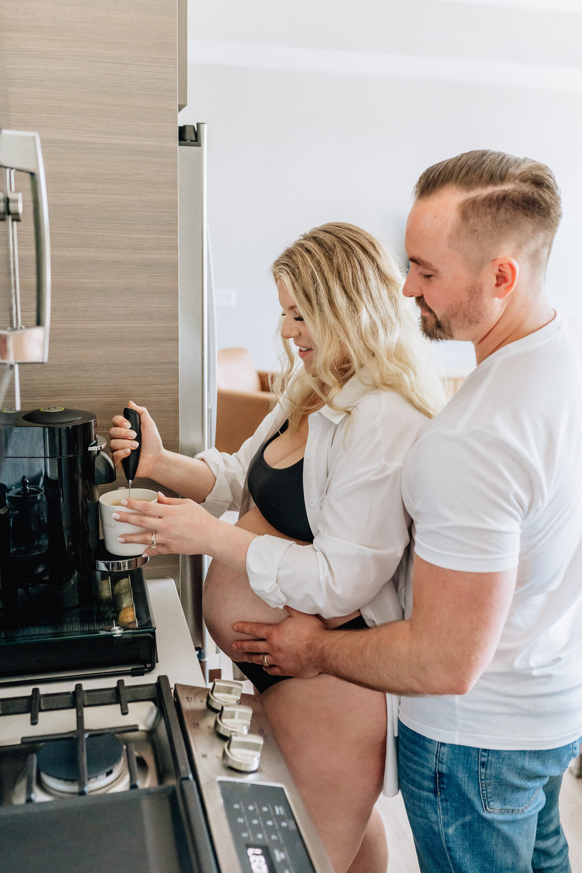 A pregnant woman in a black bra and white shirt stands in the kitchen, making coffee with a frother while her partner stands behind her, holding her belly with a loving smile.