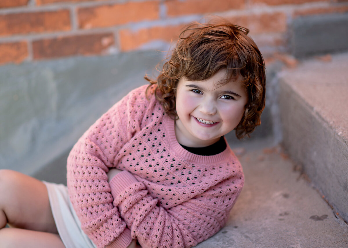 Child in a pink sweater sitting on concrete steps by a brick wall near her Iowa Milestone Photographer’s studio