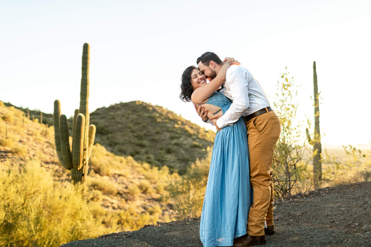 husband and wife, owner of Cactus & Pine Photography, hugging in a desert view at sunset