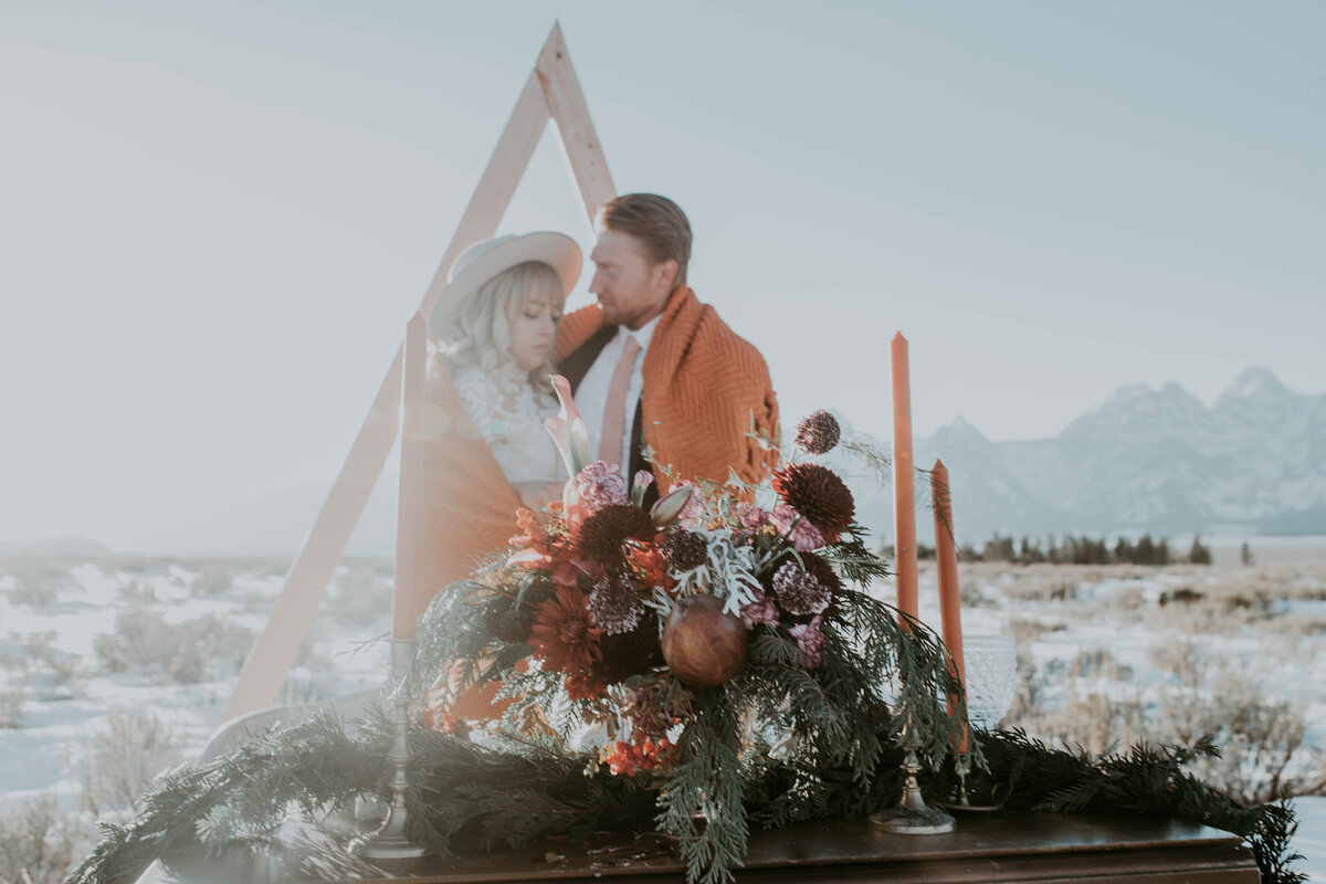 bride and groom in a winter elopement scene with a burnt orange blanket wrapped around the couples as they stand next to a triangle wedding arch and a large floral wedding centerpiece on a wood table captured by Idaho Falls wedding photographer