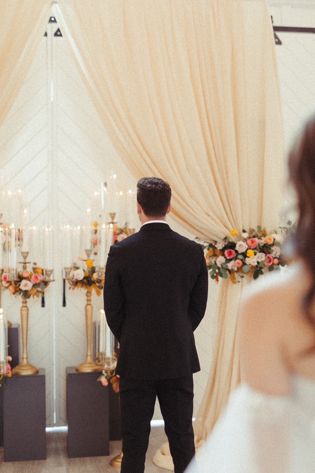 Back image of a bride wearing a white wedding gown looking at groom wearing a black tuxedo standing at the altar.