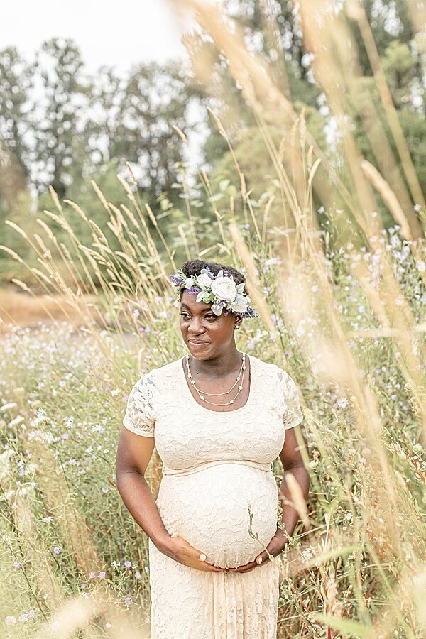 Pregnant woman in light beige lace dress wearing a floral crown holding belly and surrounded by tall grass at outdoor Oregon Maternity photo session.
