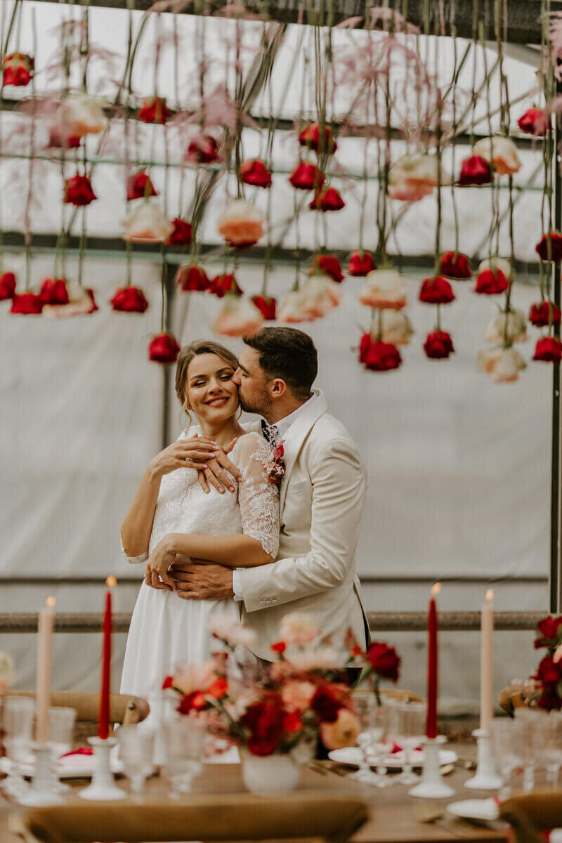 Mariés s'enlaçant dans un décor de fleurs suspendus et derrière une table en bois décorée de bougies et de fleurs, posant pour un workshop photographie de mariage.