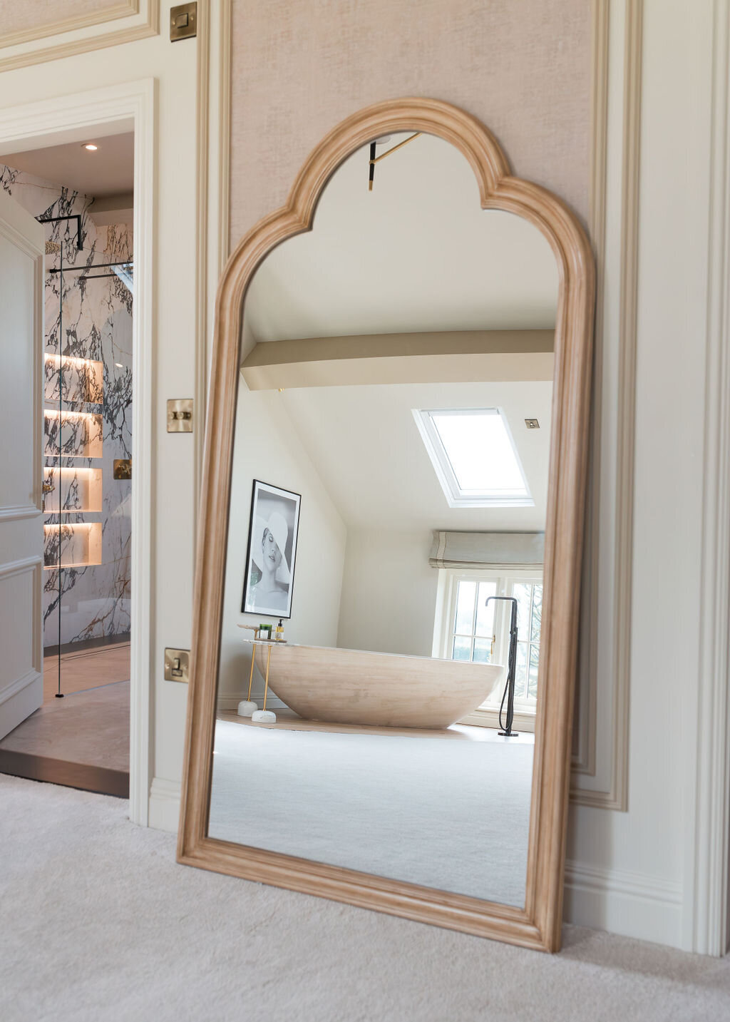 A large, ornate mirror with a wooden frame rests against a white wall in a bright room. The mirror reflects a cozy space with a freestanding bathtub under a skylight. The room exudes a serene, luxurious ambiance.