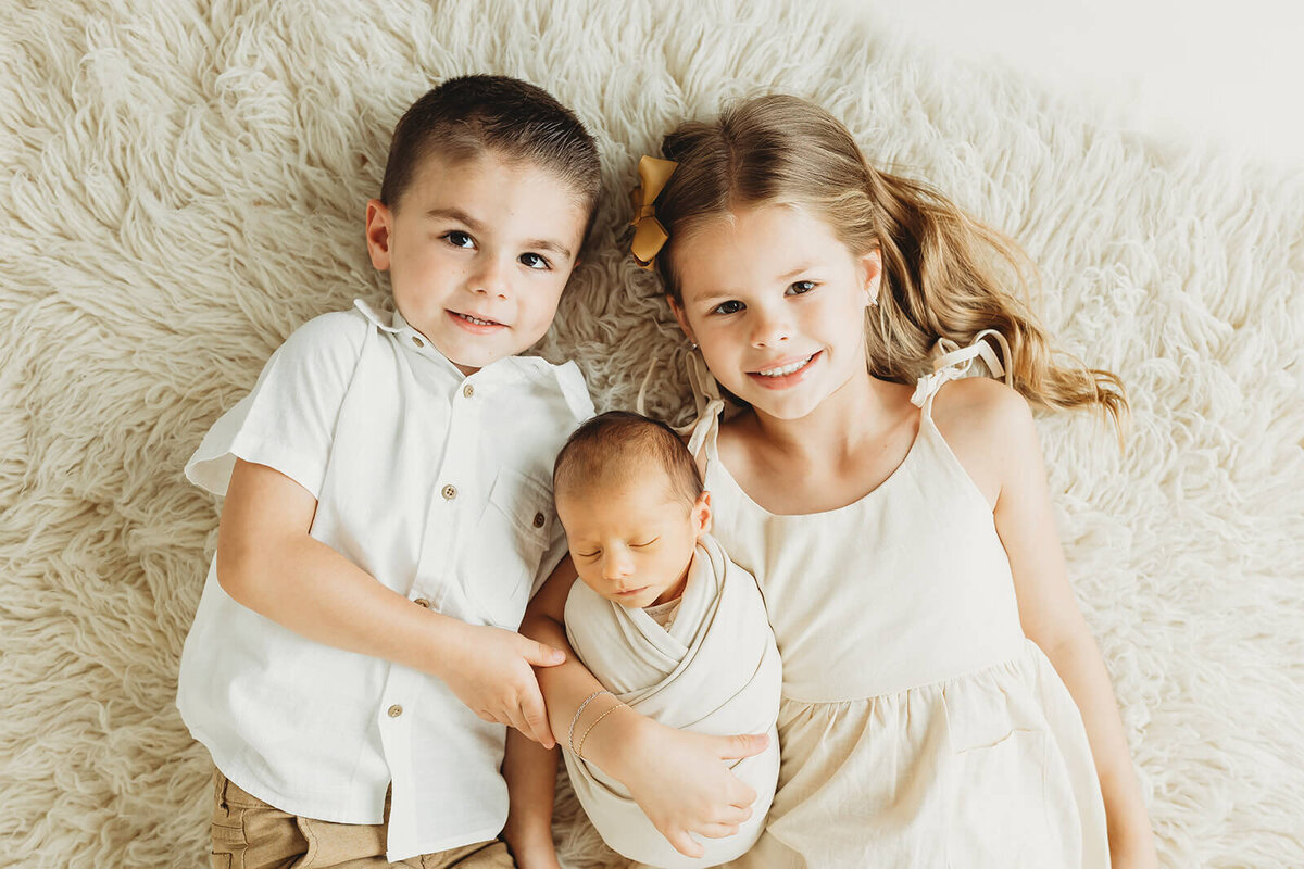 brother and sister laying on a furry rug looking up at the camera smiling holding their newborn brother