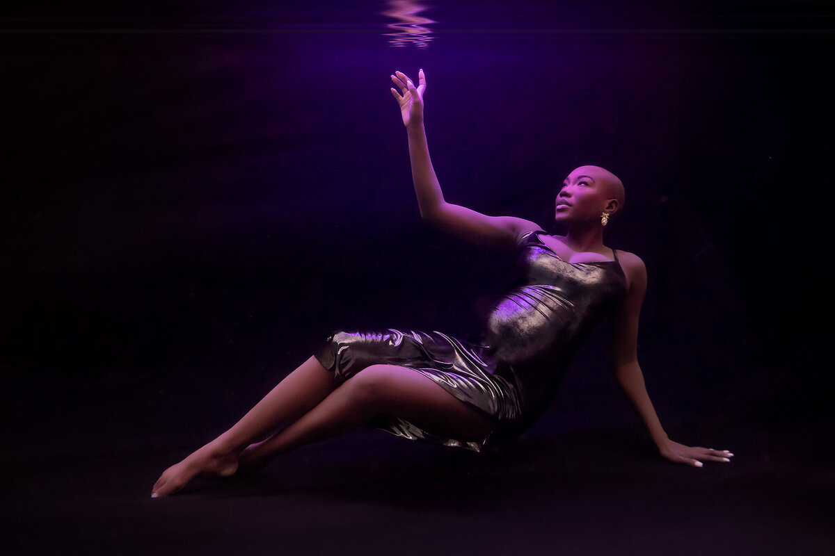 A captivating underwater maternity photo featuring a woman in a sleek, metallic dress, gracefully reaching toward the water's surface with a soft, ethereal purple glow enveloping the scene. The dramatic contrast of dark surroundings highlights the elegance and strength of the subject. This underwater maternity photoshoot beautifully showcases the artistry of  Dallas underwater pregnancy photographer Lindsay Walden.