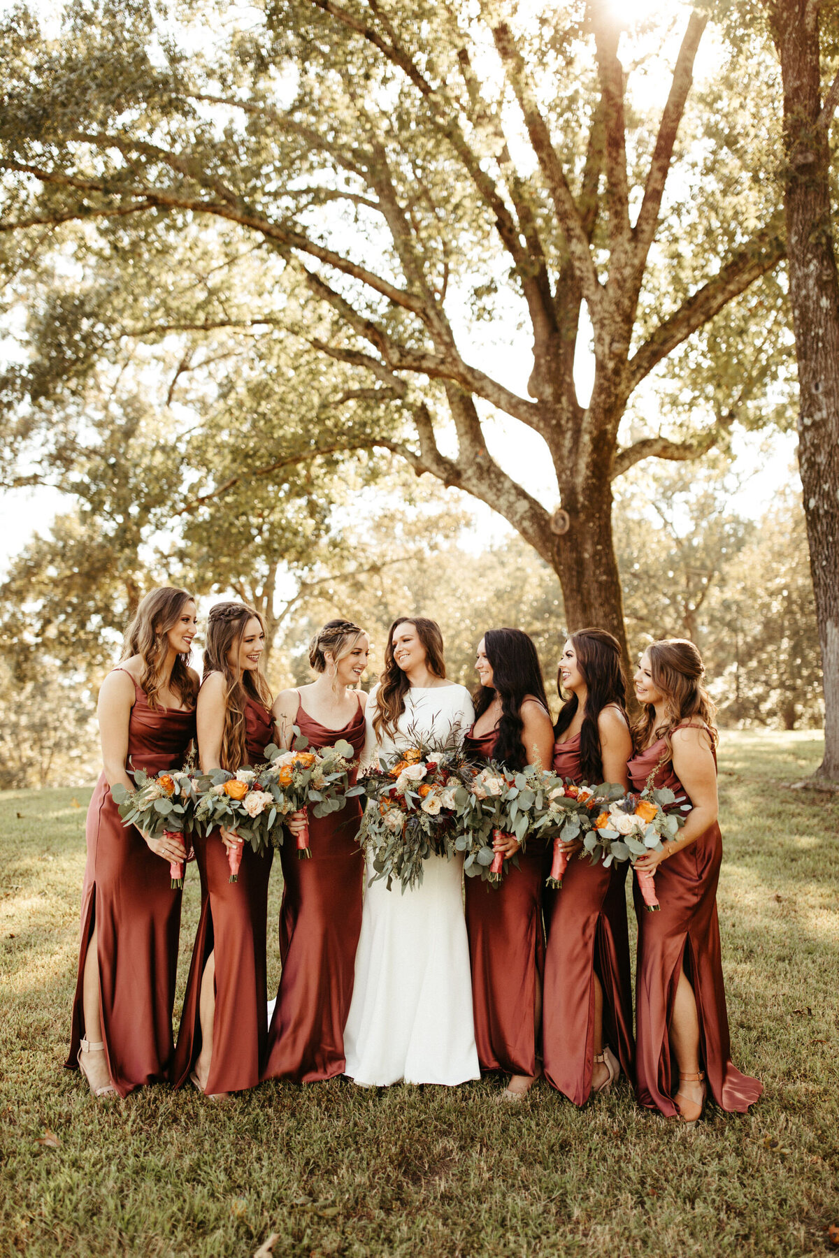 Bride with her six bridesmaids in silk sheath dresses all holding large floral bouquets with greenery and standing under a tree