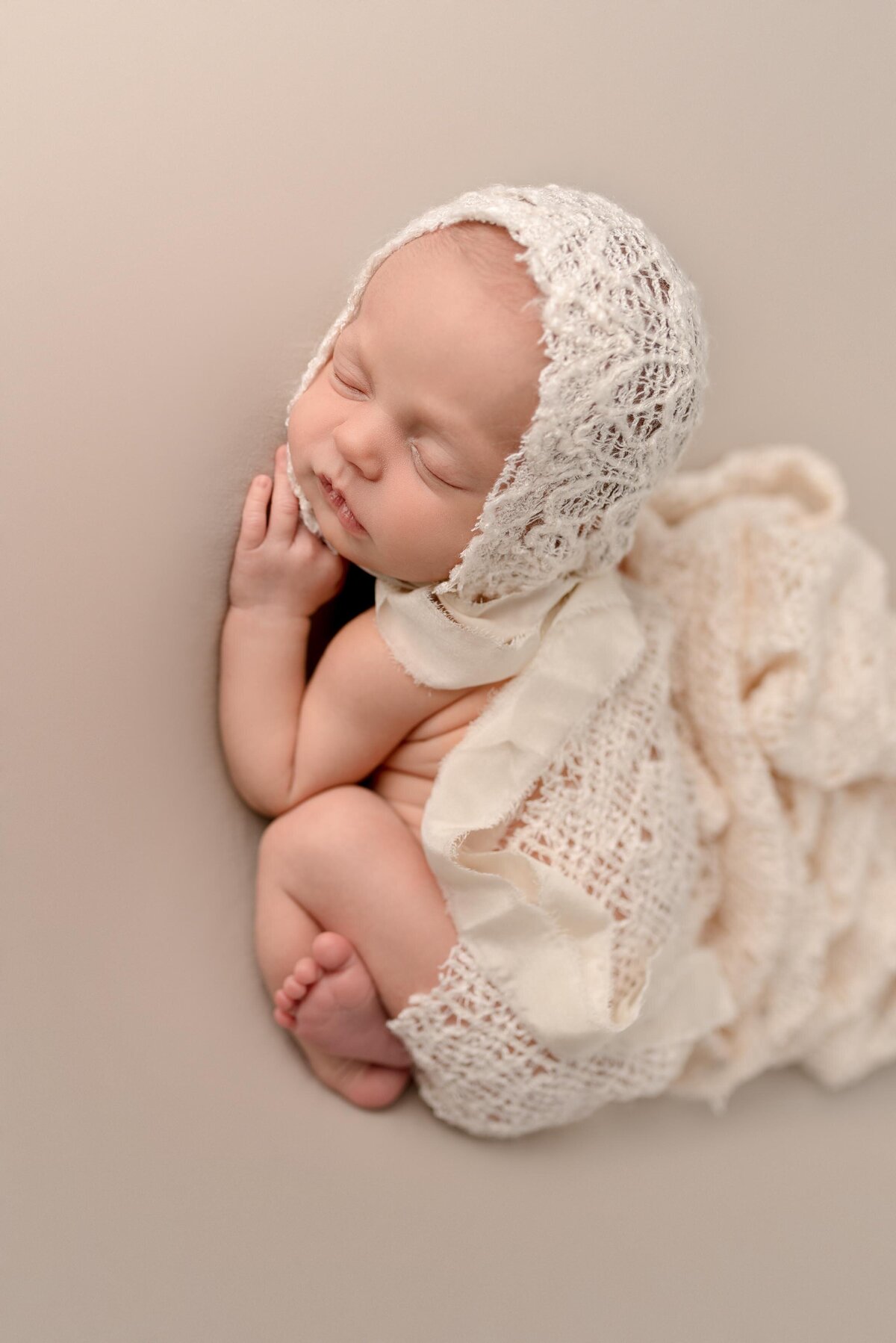 Newborn baby girl laying on her tummy in the bum up position wearing a lace bonnet and lace wrap laying over back.