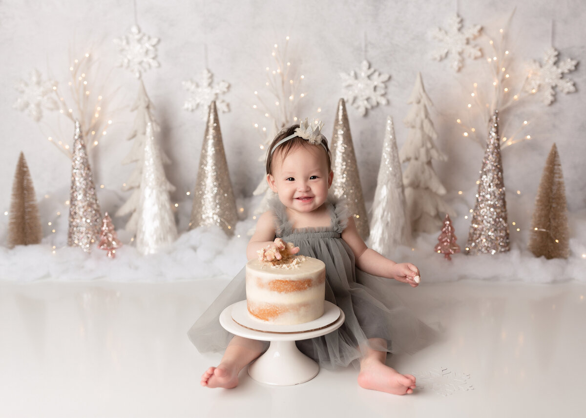 Winter Onderland themed cake smash at top West Palm Beach, FL newborn and cake smash photographer. Asian baby girl is wearing a grey tulle dress, touching the cake for the first time. She is smiling at the camera. In the background there are silver and white trees, snowflakes, and faux snow.