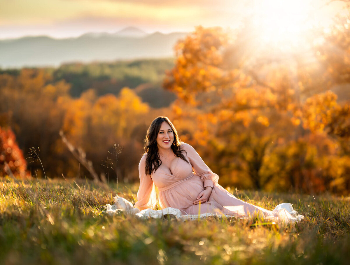 A beautiful mama to be cradles her belly while sitting on a white blanket in the grass with mountains behind her