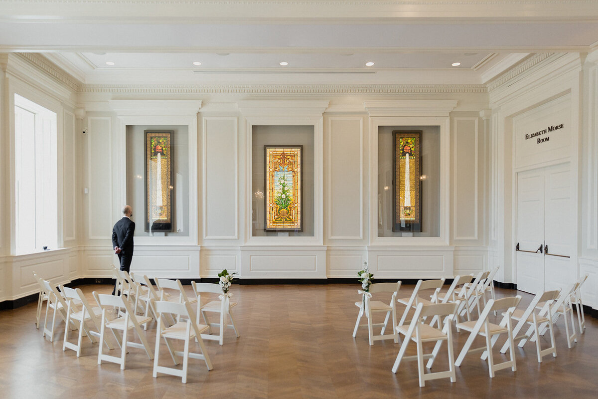 groom standing in ceremony space looking at glass window. wedding guest chairs surround him