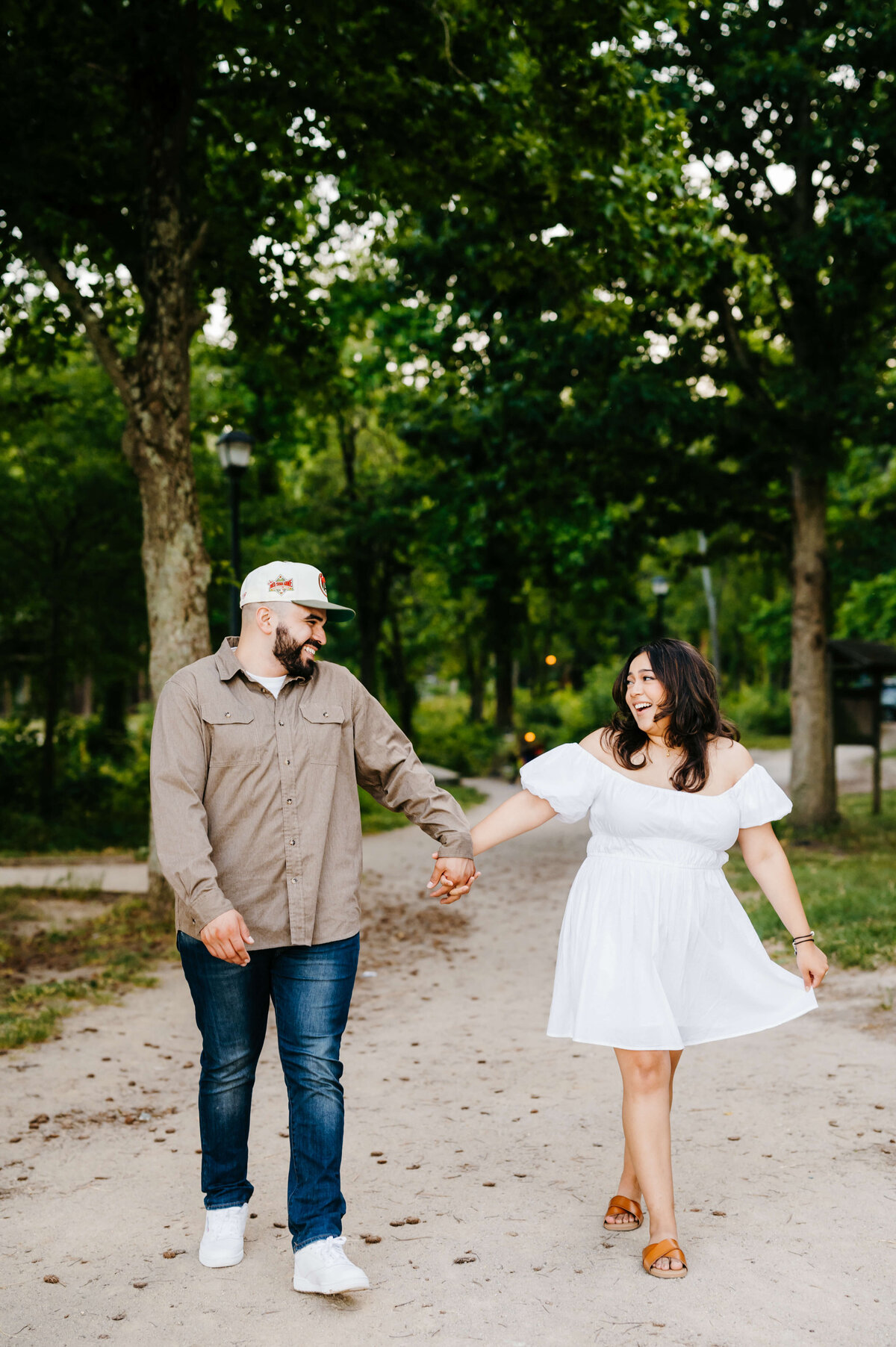 beach engagement photos with man and woman walking down a sandy path with trees on either side of the path with the man and woman holding hands and smiling