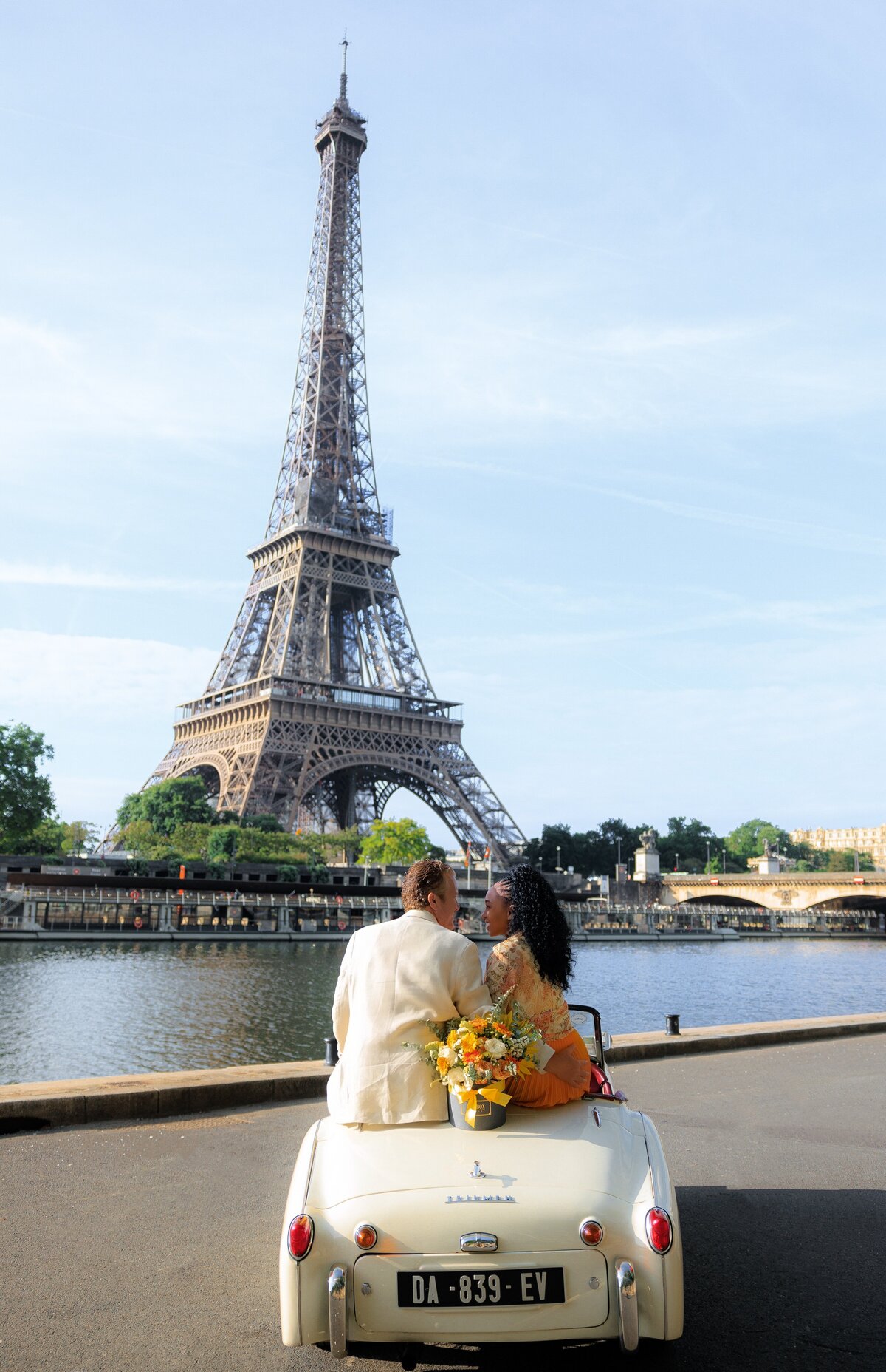 Stunning couple sit on the back of a vintage convertible car on the edge of the Seine with a view of the Eiffel Tower