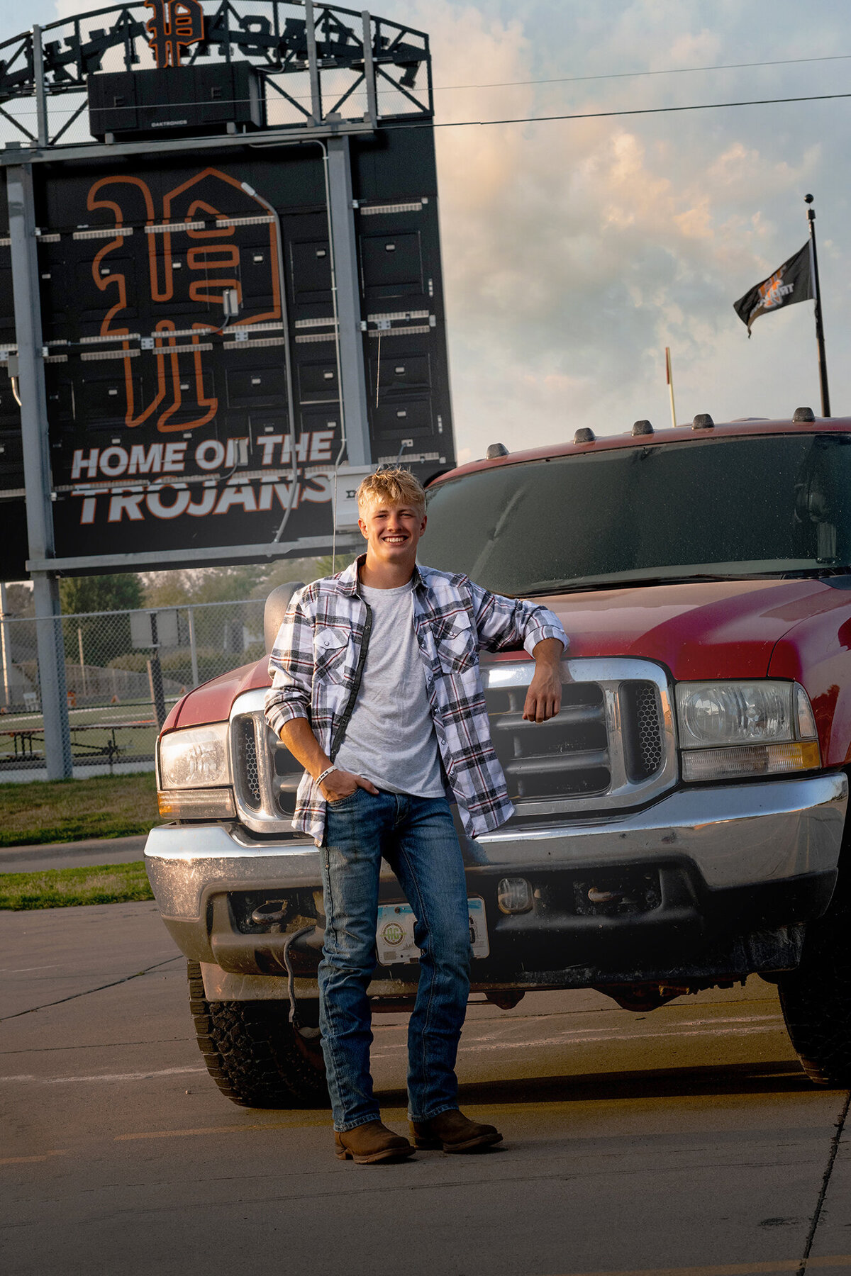 A man leans on his big red truck in the stadium parking lot at sunset