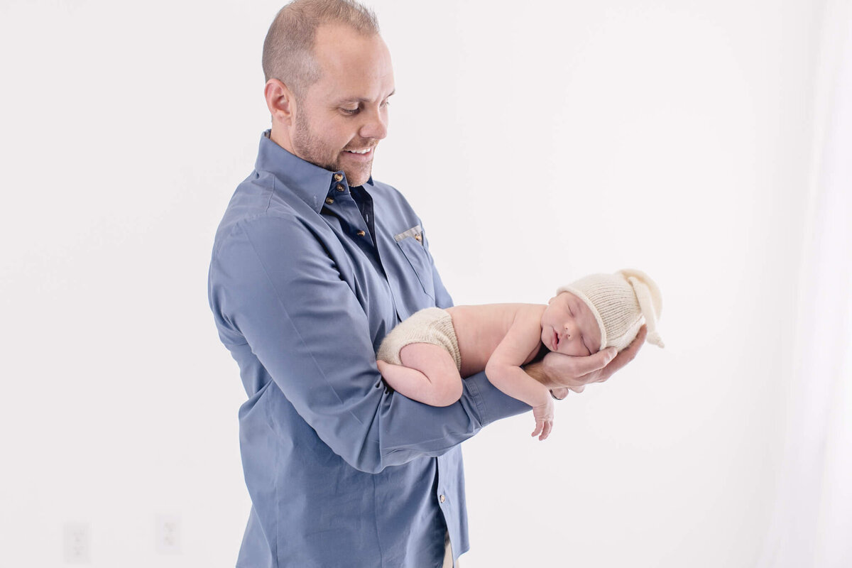 A man in a blue shirt gently holds a newborn dressed in a cream-colored knit hat and diaper against a white background. He smiles softly, looking down at the baby.