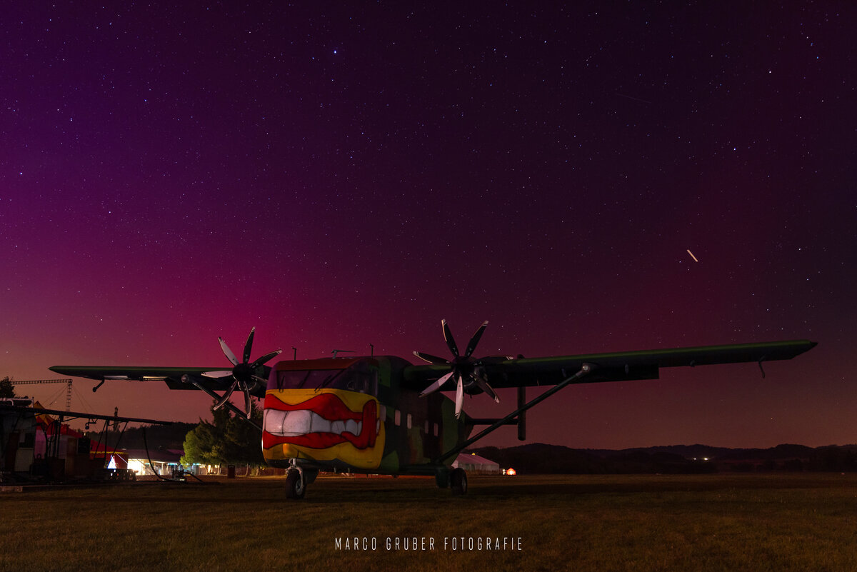 Das Bild zeigt die Pink Skyvan am Boden mit Nordlichtern. Der Himmel ist gelb, pink und violett