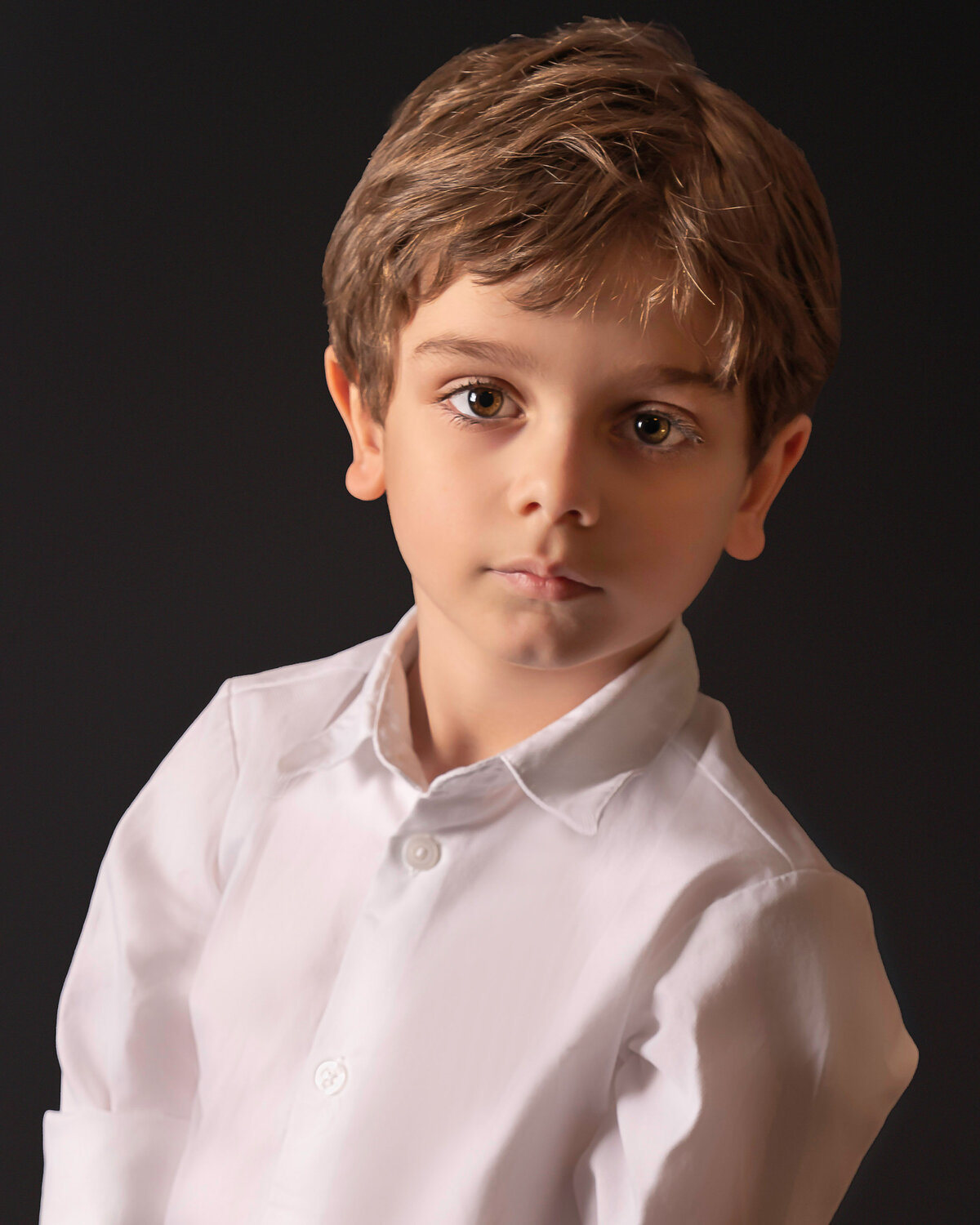 Little boy poses for studio portrait. He is wearing a white dress shirt and looking intently at the camera, no smile. Captured by Liza Nicole Photography, Philadelphia and Main Line family photographer.