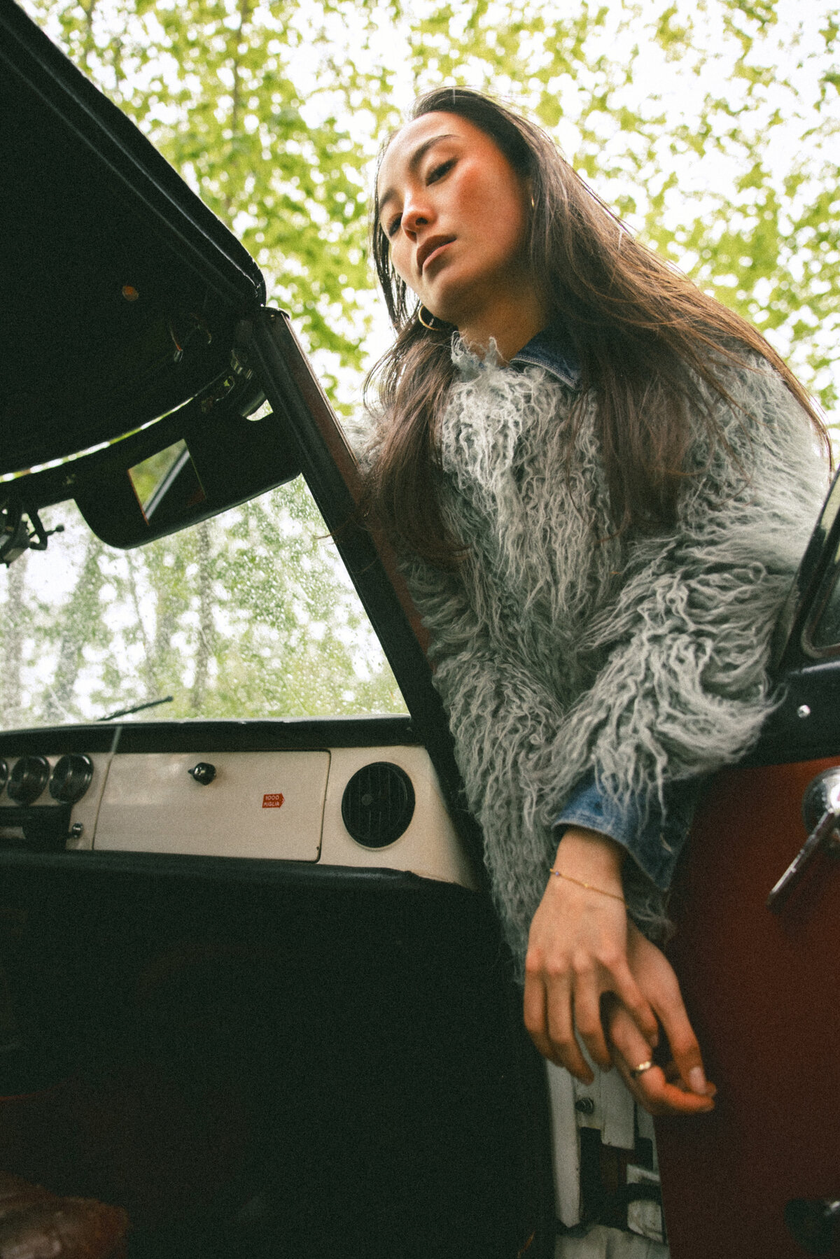 The model leans out of the car door, looking down with a serene expression. The lush greenery in the background contrasts beautifully with the structured look of her fur coat and denim, blending nature with fashion. Photographed by Bo Eppink.