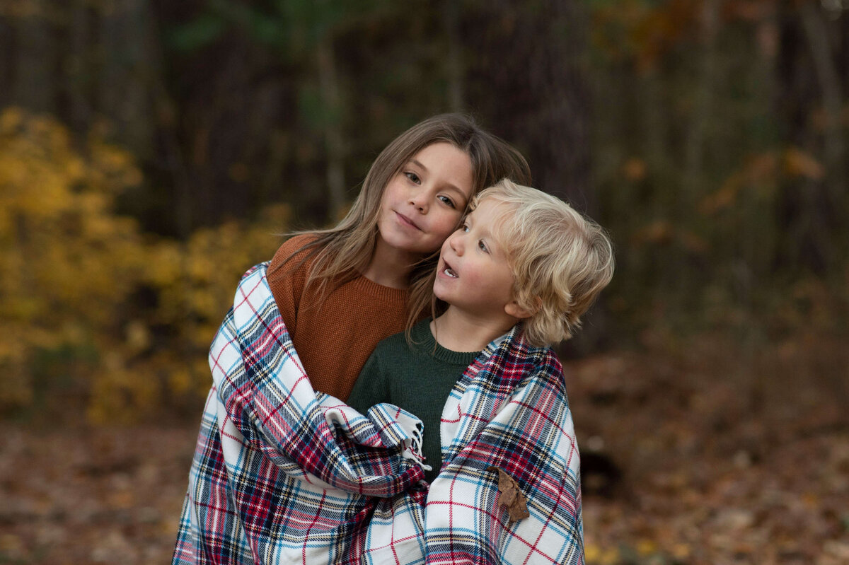 Two kids with a plaid blanket draped across their shoulders smile, one at the camera and one up at the sky.