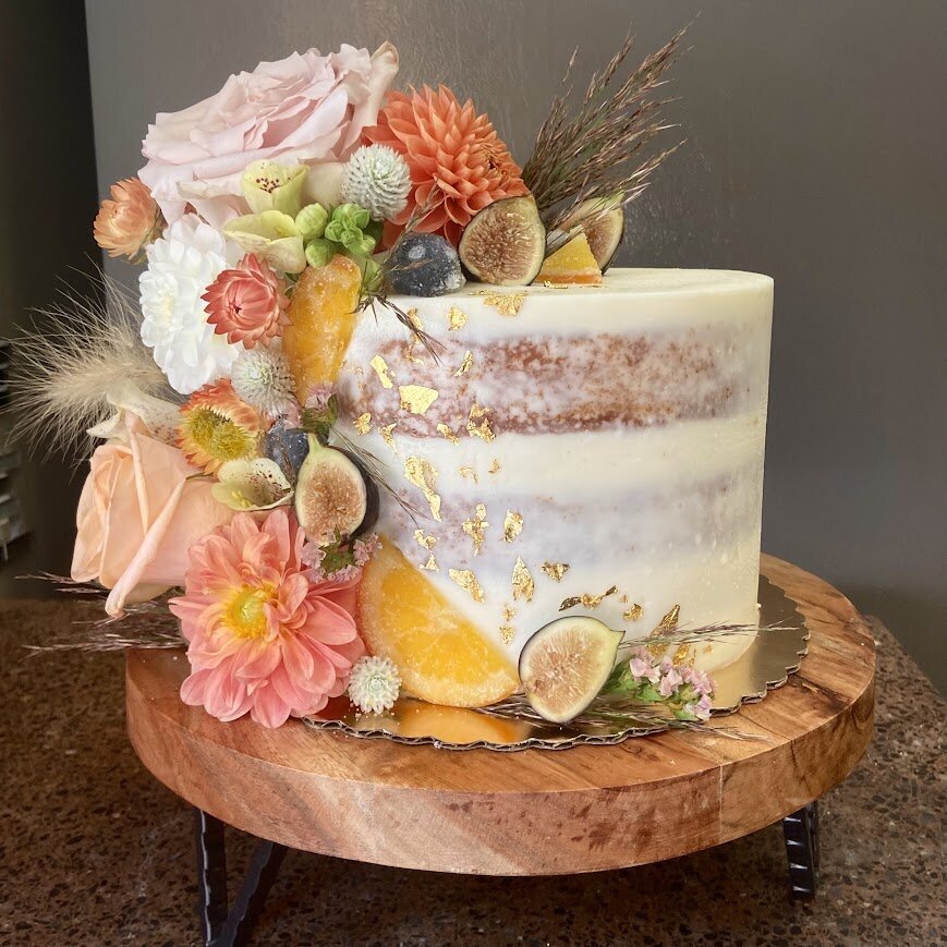 A naked style cake sports a cascade of fresh flowers in shades of pink, with fresh figs, candied oranges, and gold leaf.