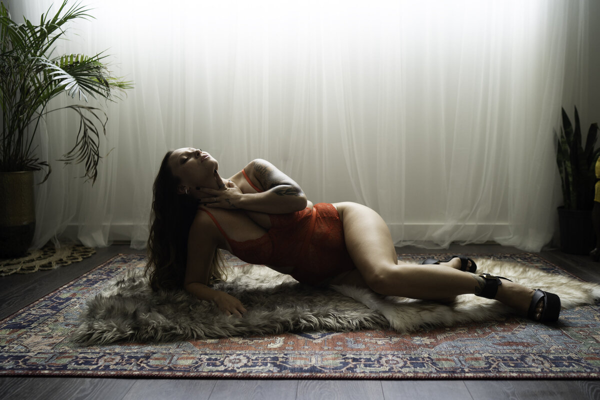 woman in red laying down on layered rugs, with plants and beautiful glowing light in a boudoir studio in Detroit.