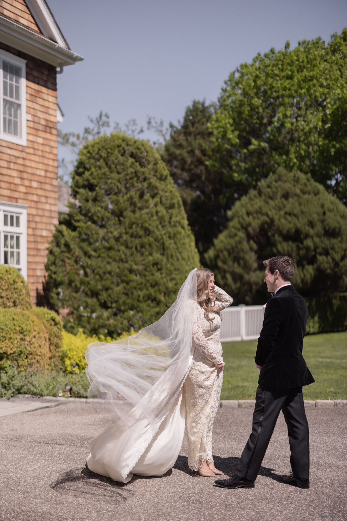 Bride and groom share a private first look outside a shingle-style estate in the Hamptons, with the bride’s veil flowing in the wind and the couple smiling at each other.