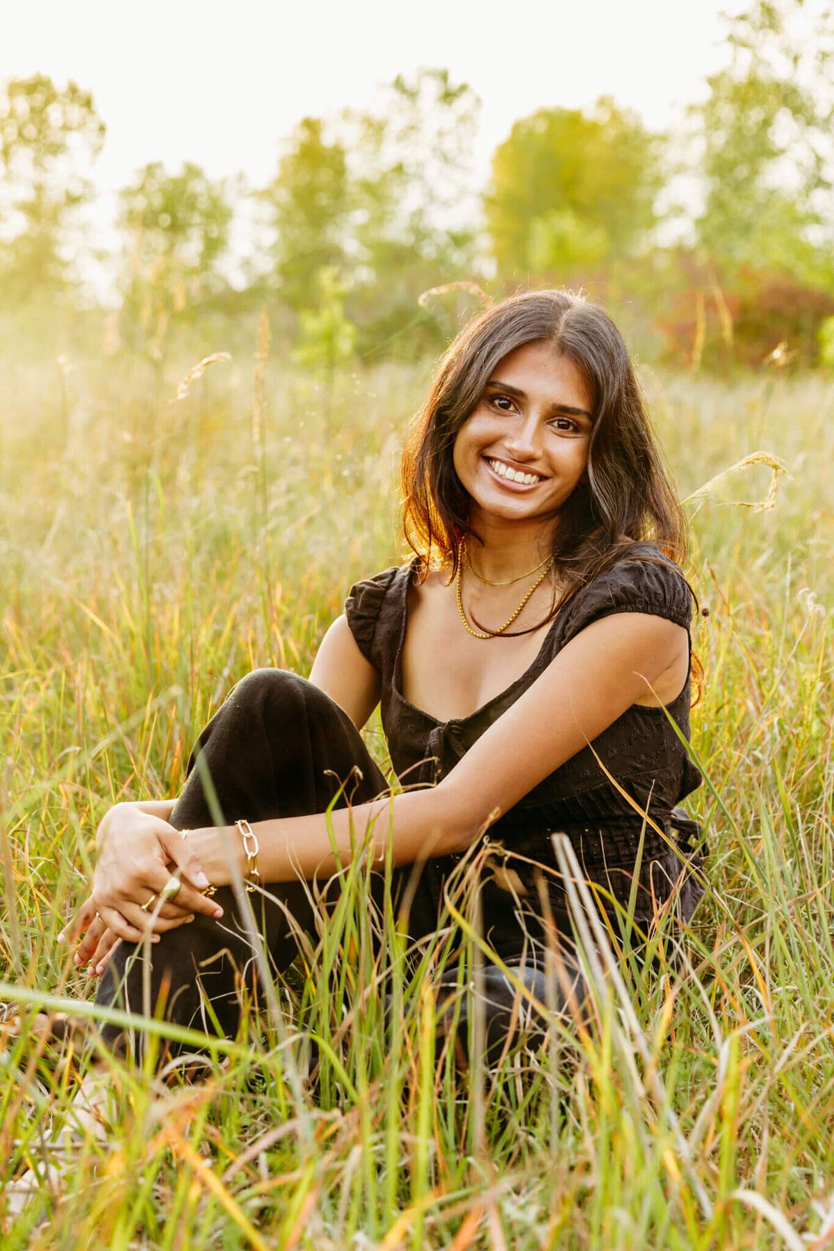 stunning teen girl sitting in some tall grass and holding her knee as she smiles.