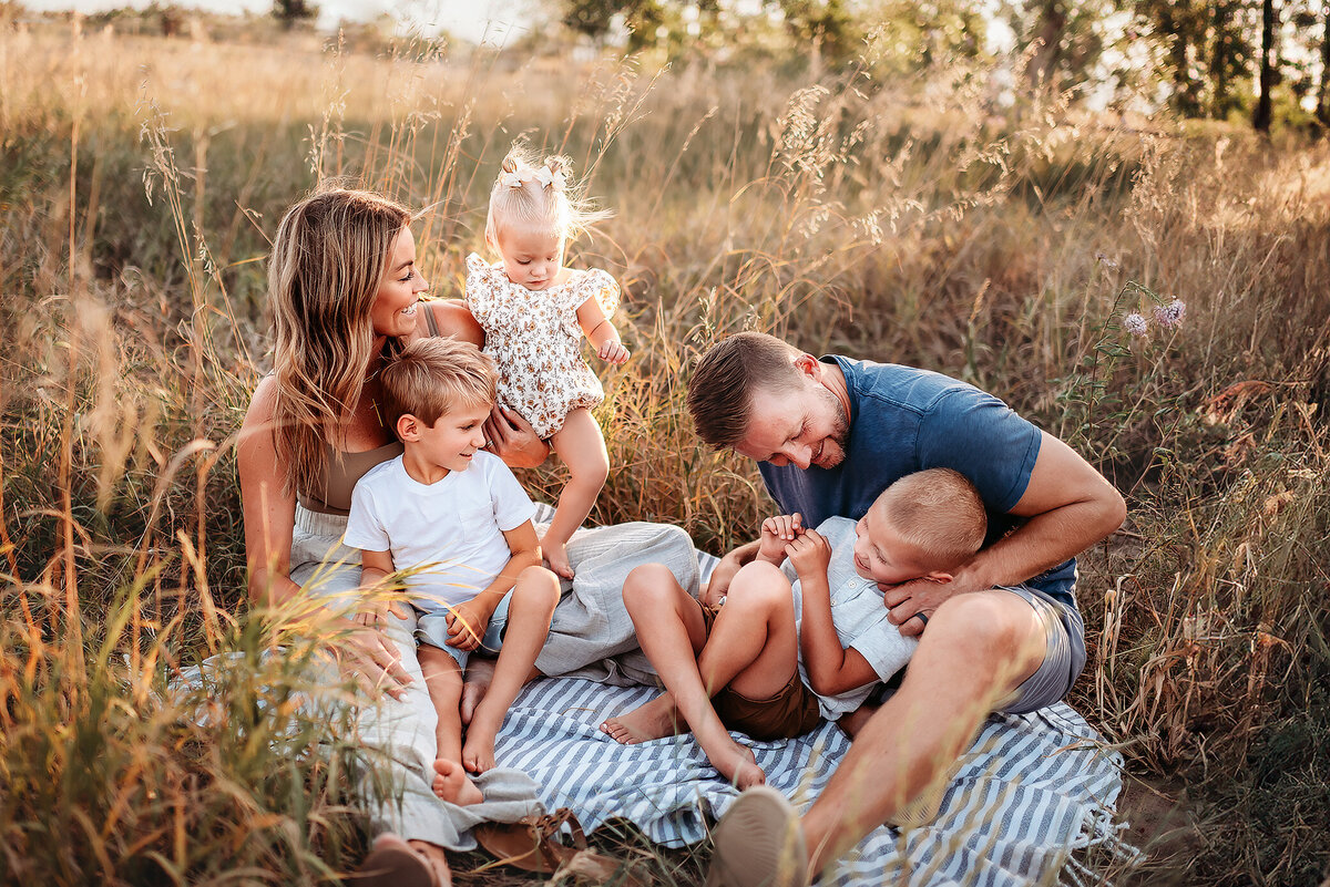family of five on a blanket in a field with dad tickling son