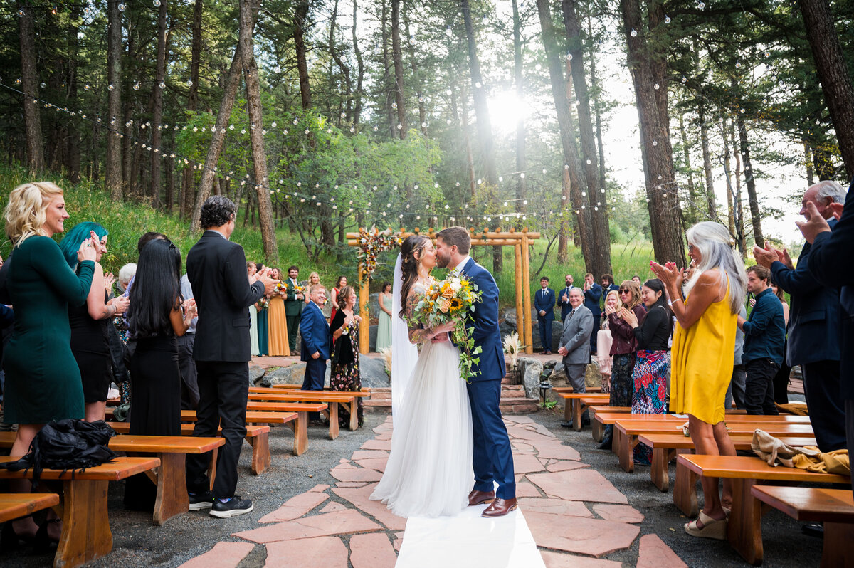 A wide angle shot of a bride and groom sharing a kiss at the end of the aisle at their outdoor ceremony at The Pines at Genesee.