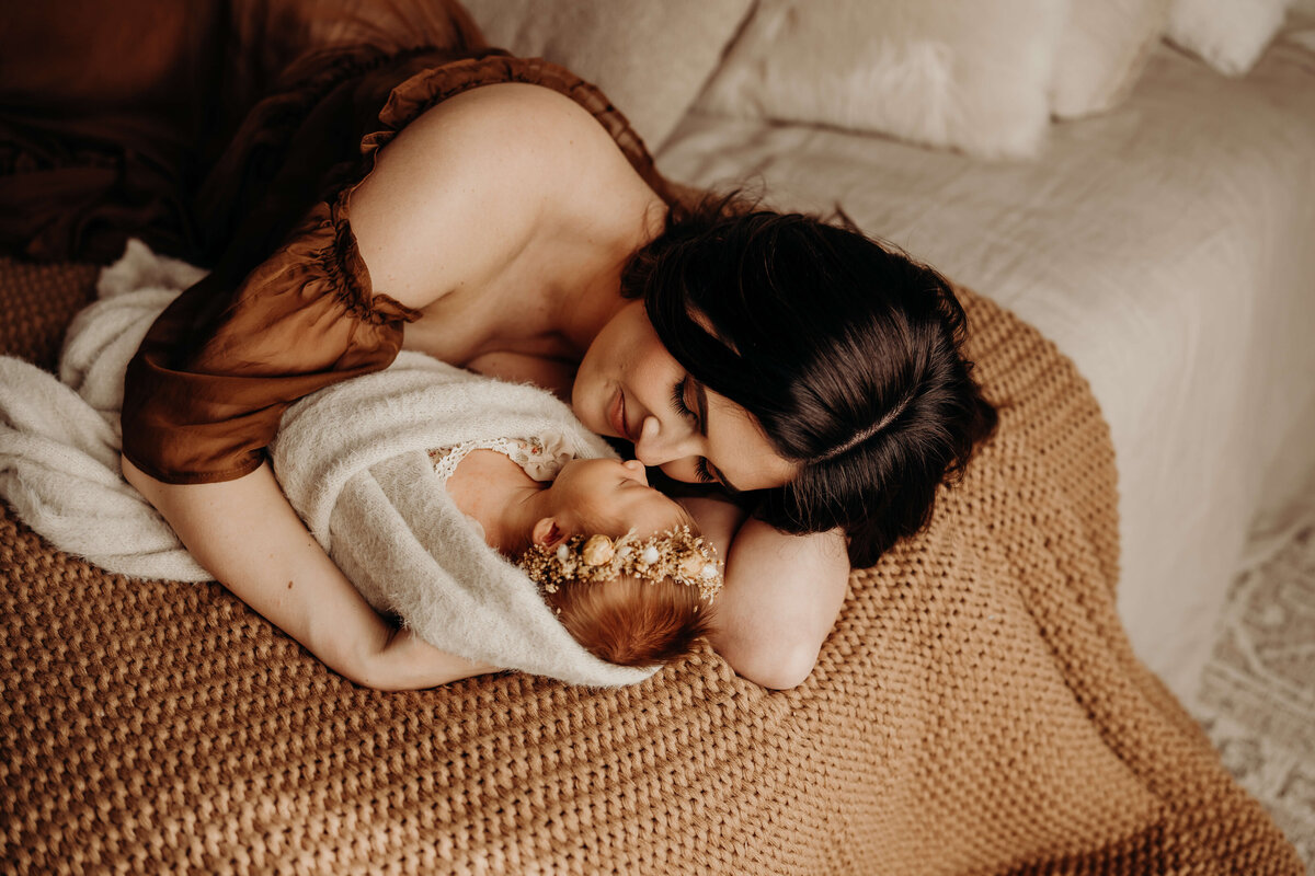 mother lays on bed in brown gown holding her newborn