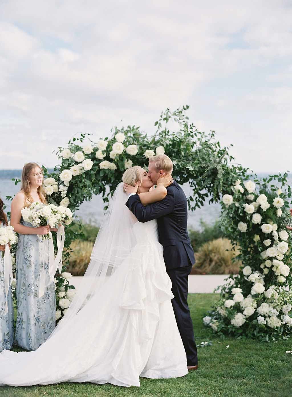 bride and groom kissing at wedding in front of greenery circle arch with white flowers