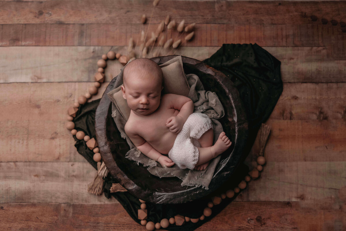A newborn boy posed in a brown bucket by Mobile, AL newborn photographer. The baby boy is laying comfortably on a pillow and green blanket inside the bowl. The backdrop is farm house wood floor.
