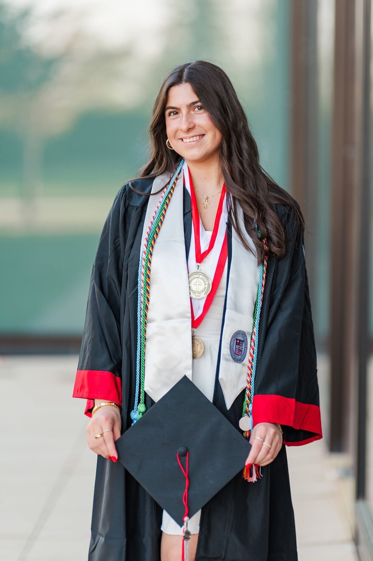 Sacred Heart University graduate wearing a white dress and her full cap and gown leans against a window.